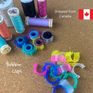 May include: A collection of colorful bobbin clips for organizing sewing thread spools. The clips are made of clear plastic and come in a variety of colors, including pink, blue, green, yellow, and orange. The clips are arranged in a pile on a brown surface. The image also includes several spools of thread in various colors, including white, pink, blue, and green.