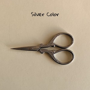 May include: Silver-colored small scissors with an ornate design. The scissors have a pointed tip and a small loop on the handle.