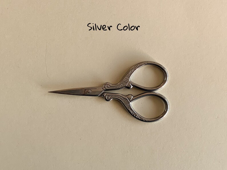 May include: Silver-colored small scissors with an ornate design. The scissors have a pointed tip and a small loop on the handle.