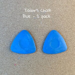 May include: Two blue tailor's chalk triangles, each with a round center, on a brown background. The text "Tailor's Chalk Blue - 2 Pack" is written in white above the chalk.