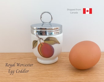 May include: A white ceramic egg coddler with a silver lid and a plum design. The coddler is on a wooden surface next to a brown egg. The text "Royal Worcester Egg Coddler" is visible below the coddler.