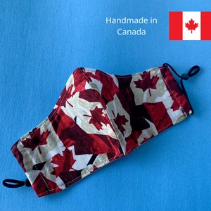 May include: A red, white, and beige face mask with a Canadian maple leaf pattern. The mask has black elastic ear loops and the text "Handmade in Canada" is visible in the background.