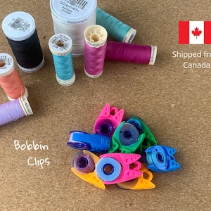 May include: A collection of colorful bobbin clips for sewing, with a variety of colors including pink, blue, green, and purple. The clips are designed to hold spools of thread in place.