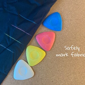 May include: Four triangular fabric marking tools in blue, red, yellow, and white. The tools are on a corkboard surface with the text "safely mark fabric" written in white.