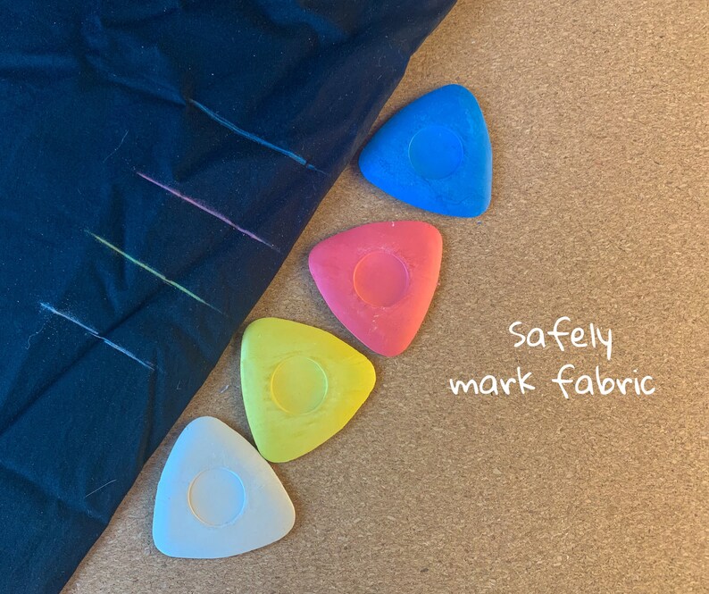 May include: Four triangular fabric marking tools in blue, red, yellow, and white. The tools are on a corkboard surface with the text "safely mark fabric" written in white.