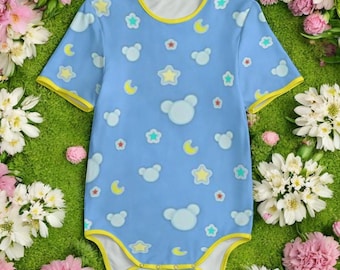 May include: A light blue baby bodysuit with a pattern of white bear faces, stars, and crescent moons. The bodysuit has yellow trim around the neckline, sleeves, and legs. The background is a bed of green grass and flowers.