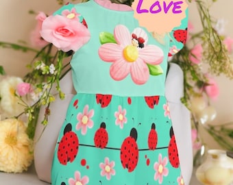 May include: A turquoise dress with a pink flower and ladybug print. The dress has short sleeves and a ruffled neckline. The text "LOVE" is written on a peach-colored circle.