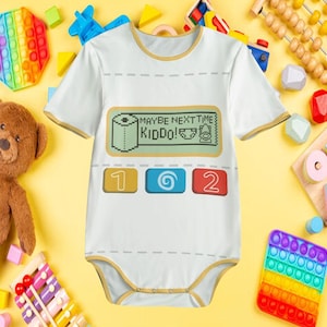 May include: A white short-sleeve baby bodysuit with gold trim. The front features a graphic of a digital screen with the text MAYBE NEXT TIME KIDDO! and icons of a toilet paper roll, a diaper, and a potty, plus three buttons labeled 1, 2.