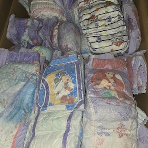 May include: A collection of disposable diapers featuring various colorful cartoon character prints including Mickey Mouse, Ariel from The Little Mermaid, and Paw Patrol characters. The diapers are white with purple, blue, and pink accents.