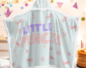 May include: A light blue hooded towel with the text "LITTLE PRINCESS" in purple and pink. The towel features pink bows and stars, with a white fluffy trim. A crown graphic is also visible.