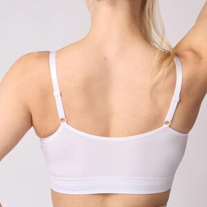 Op de afbeelding: Een witte bralette met een diepe ruguitsnijding, dunne verstelbare schouderbandjes met metalen schuifjes en een brede elastische band onder de buste. Het kledingstuk is gemaakt van een gladde, effen witte stof met strakke randen.
