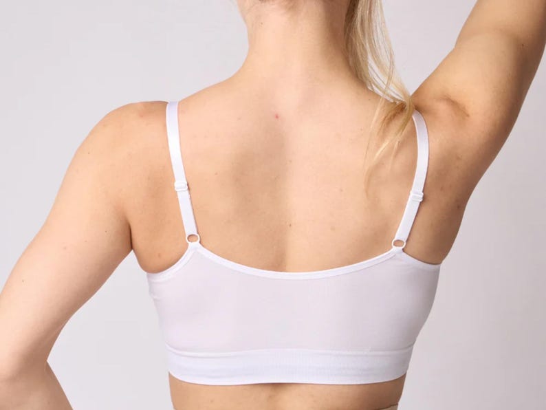 Op de afbeelding: Een witte bralette met een diepe ruguitsnijding, dunne verstelbare schouderbandjes met metalen schuifjes en een brede elastische band onder de buste. Het kledingstuk is gemaakt van een gladde, effen witte stof met strakke randen.