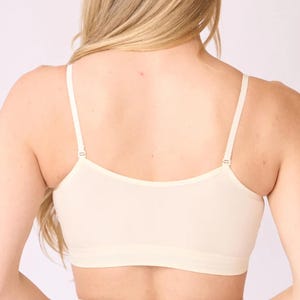 Op de afbeelding: Een beige naadloze bralette met een diepe ruguitsnijding, dunne verstelbare schouderbandjes met metalen schuifjes en een brede elastische band onder de buste. Het kledingstuk is gemaakt van een effen, gladde stof met een nette afwerking.