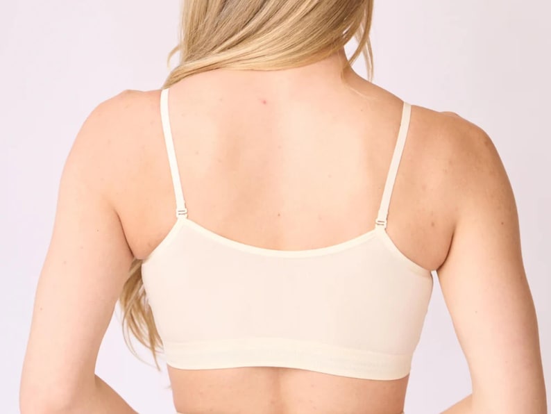 Op de afbeelding: Een beige naadloze bralette met een diepe ruguitsnijding, dunne verstelbare schouderbandjes met metalen schuifjes en een brede elastische band onder de buste. Het kledingstuk is gemaakt van een effen, gladde stof met een nette afwerking.
