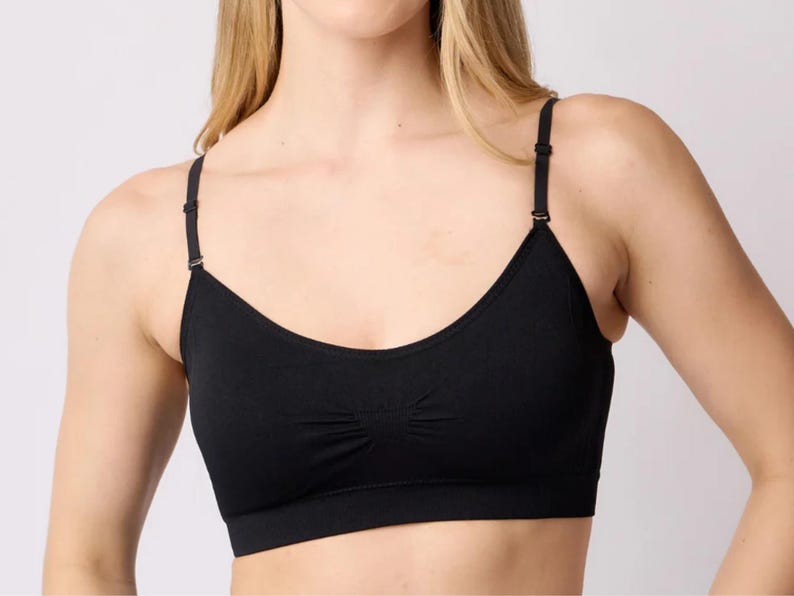 Op de afbeelding: Een effen zwarte bralette met een ronde halslijn, dunne verstelbare schouderbandjes met metalen schuifjes en een subtiel geplooid detail in het midden van de borst. Het kledingstuk heeft een brede, platte elastische band aan de onderkant.