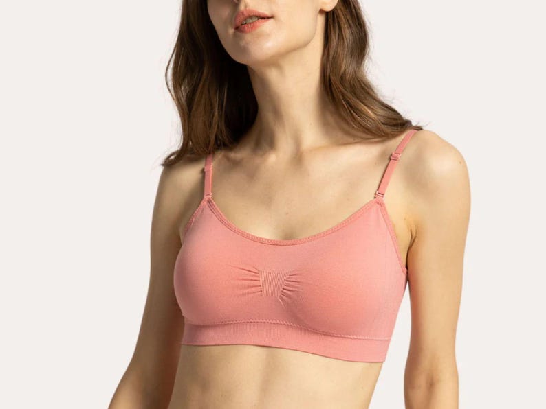 Op de afbeelding: Een naadloze bralette in een effen koraalkleur met dunne, verstelbare schouderbandjes, een ronde halslijn en een brede band onder de buste. De stof heeft een subtiele ribbeltextuur met een klein geplooid detail in het midden van de borst.