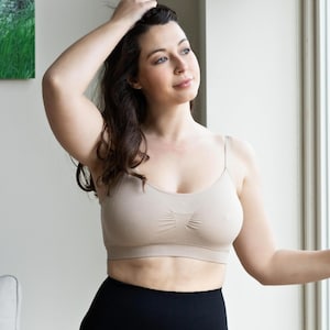 May include: A beige seamless bralette featuring thin spaghetti straps, a scoop neckline, and a ribbed center detail between the cups. The garment is constructed from a smooth, stretchable fabric with a wide band along the bottom hem.