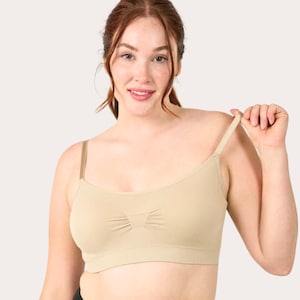 May include: A beige seamless bralette featuring thin adjustable shoulder straps, a scoop neckline, and a subtle ruched detail at the center of the chest. The garment has a wide, ribbed underband for support and a smooth, solid-colored fabric finish.