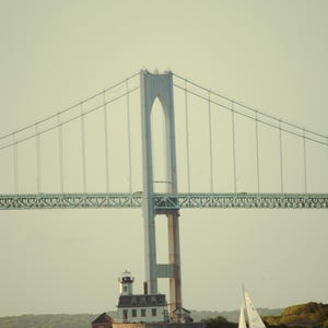 Op de afbeelding: Een grote hangbrug met een hoge centrale toren overspant een watermassa. Onder de brug staat een witte vuurtoren op een rotsachtig eiland en een witte zeilboot met één mast vaart op het water nabij de oever.