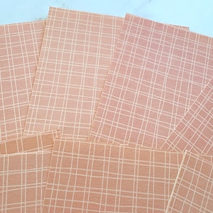 Peut inclure: Une pile de feuilles de papier couleur pêche avec un motif quadrillé blanc. Les feuilles sont disposées en éventail, certaines se chevauchant. L'arrière-plan est une surface bleu clair.