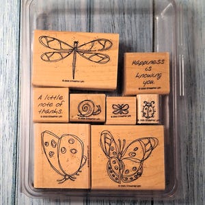 May include: A collection of wooden stamps with various insect and text designs. The stamps include a dragonfly, butterflies, a ladybug, a snail, and the phrases "A little note of thanks" and "Happiness is knowing you."