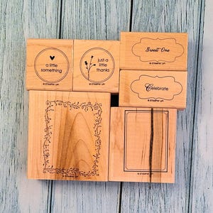 May include: A collection of wooden stamps with various designs and text. The stamps include phrases like "Sweet One" and "Celebrate," along with decorative elements such as a heart, flowers, and a framed border. The stamps are made of light-colored wood.