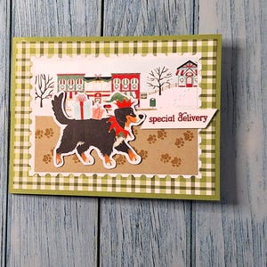 May include: A greeting card with a festive design. The card features a black and brown dog wearing a red collar and a green hat, walking in front of a winter village scene. The words "special delivery" are written on a white banner. The card has a green and white checkered border.