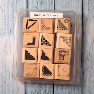 May include: A clear plastic storage case containing twelve square wooden rubber stamps labeled Creative Corners. Each stamp features a unique black ink corner design, including geometric patterns, floral swirls, and textured borders.