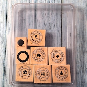 May include: A collection of wooden stamps with various designs and phrases, including "WELCOME LITTLE ONE," "LOVE FOR YOU," and "HAPPY HOLIDAYS." The stamps are in a clear plastic container, set against a light blue wooden background.