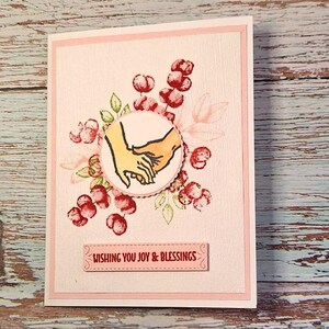 May include: A handmade greeting card with a pink border and a textured background. The card features a central illustration of two hands clasped together, surrounded by red floral elements and green leaves. The text "WISHING YOU JOY & BLESSINGS" is displayed on a pink banner.