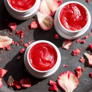May include: Three small white jars filled with a bright red lip balm. The jars are sitting on a black surface with dried rose petals scattered around them.
