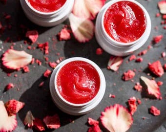 May include: Three small white jars filled with a bright red lip balm. The jars are sitting on a black surface with dried rose petals scattered around them.