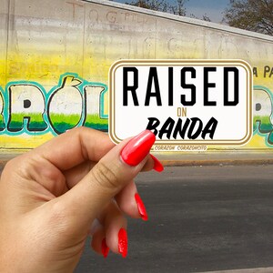 May include: A white sticker with gold trim that reads "RAISED ON BANDA" in black text. The sticker is held in a hand with red nail polish.