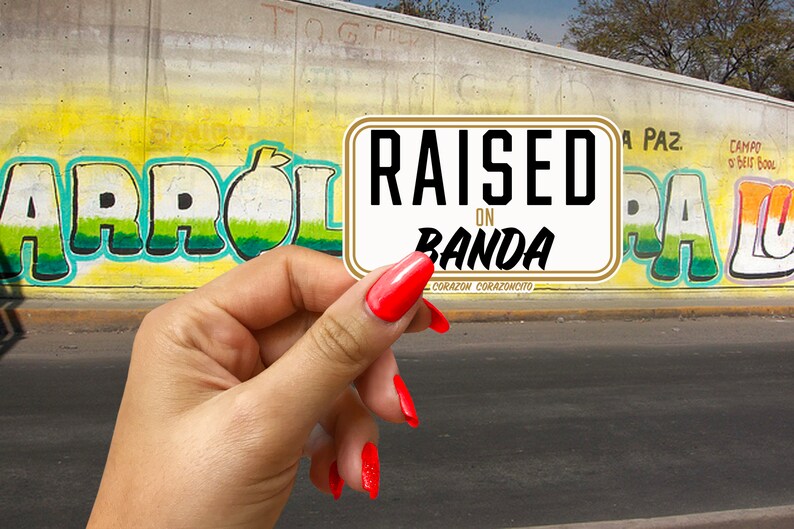 May include: A white sticker with gold trim that reads "RAISED ON BANDA" in black text. The sticker is held in a hand with red nail polish.