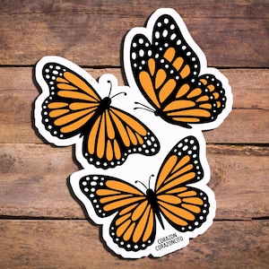 May include: Three monarch butterfly stickers with orange and black wings and white borders are displayed on a wooden surface. The stickers have a detailed design, with the text "CORAZON CORAZONCITO" on one.