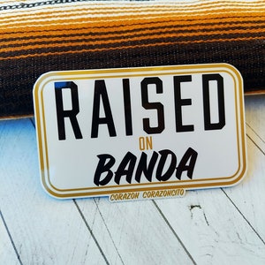 May include: White sticker with gold trim and black text that reads "RAISED ON BANDA" with the words "CORAZON CORAZONCITO" at the bottom.