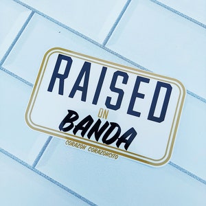 May include: A white sticker with gold trim and black text that reads "RAISED ON BANDA" with the words "CORAZON CORAZONCITO" below.
