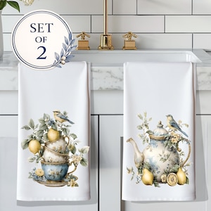May include: A set of two white kitchen towels featuring printed illustrations. One towel displays stacked teacups with lemons and a small bird. The other towel shows a floral teapot with lemons and a bird. A circular badge reads SET OF 2.