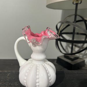 May include: A white milk glass pitcher with a pink ruffled top and a beaded design. The pitcher has a handle and a round body.