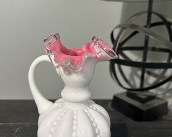 May include: A white milk glass pitcher with a pink ruffled top and a beaded design. The pitcher has a handle and a round body.