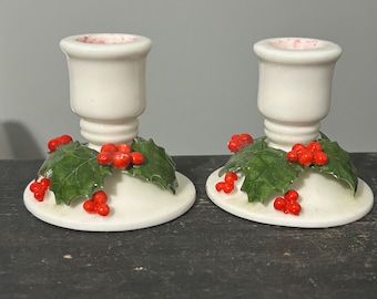 May include: Pair of white ceramic candle holders with a festive holly berry design. Each holder features a base adorned with green leaves and red berries. The candle holders are designed to hold standard-sized candles.