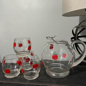 May include: A set of four clear glass tumblers and a pitcher with red apple designs. The glasses and pitcher have a rounded shape and a clear glass base.