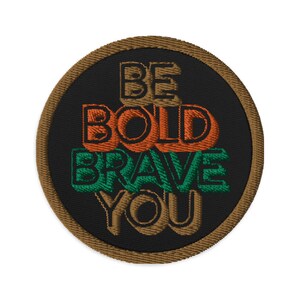 May include: A round, brown embroidered patch with the text "BE BOLD BRAVE YOU" in orange, green, and black lettering.