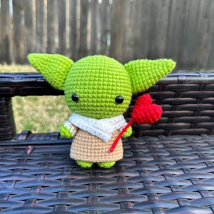 May include: A crocheted green figure with large ears, black eyes, and a tan and white outfit. The figure is holding a red heart on a red stick. The figure is sitting on a dark brown woven surface.