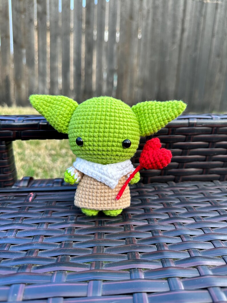 May include: A crocheted green figure with large ears, black eyes, and a tan and white outfit. The figure is holding a red heart on a red stick. The figure is sitting on a dark brown woven surface.