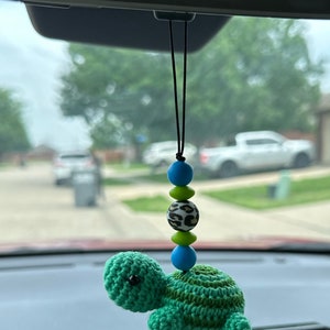 May include: A green crocheted turtle hanging from the rearview mirror of a car. The turtle is attached to a string with colorful beads.
