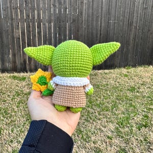 May include: A handmade green crocheted figure with large ears, wearing a tan robe with a white collar. The figure is holding a small yellow and green crocheted flower. The background is a wooden fence and grass.