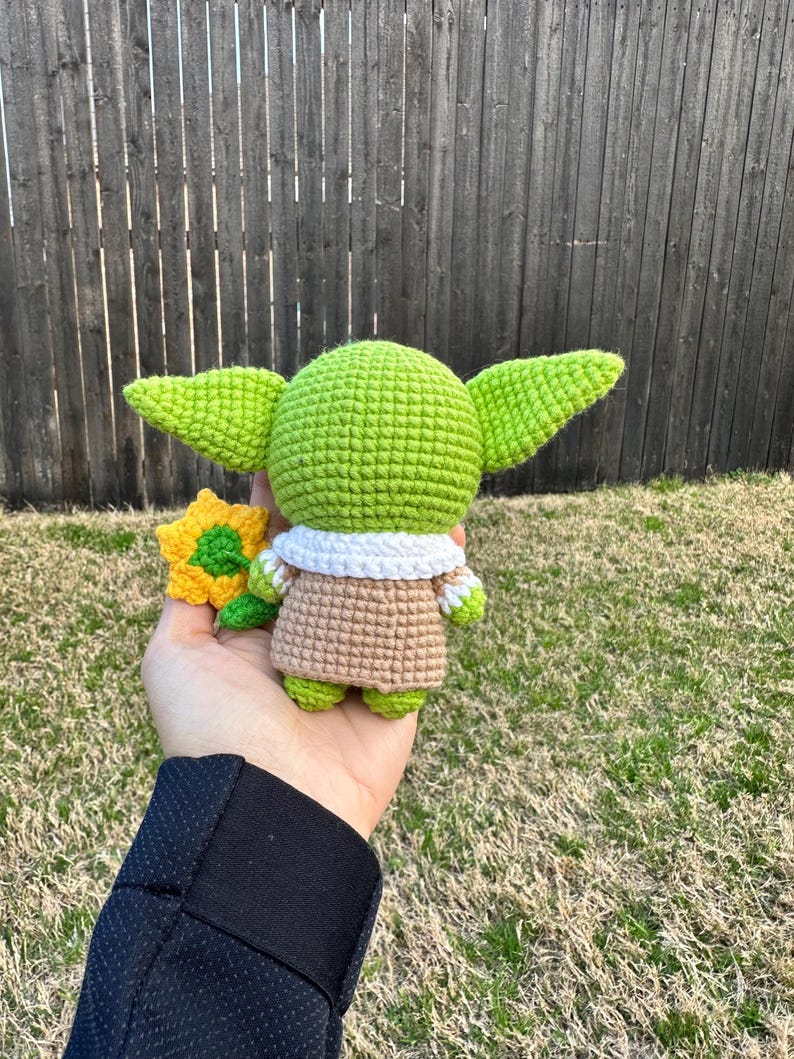 May include: A handmade green crocheted figure with large ears, wearing a tan robe with a white collar. The figure is holding a small yellow and green crocheted flower. The background is a wooden fence and grass.