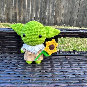 May include: A handmade green crocheted figure with large ears, black eyes, and a tan robe, holding a yellow sunflower with a green stem. The figure is sitting on a dark brown wicker bench.