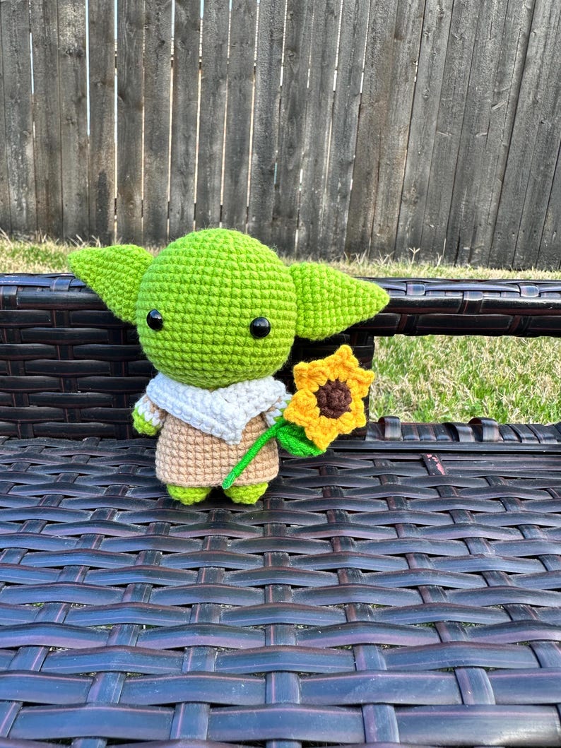 May include: A handmade green crocheted figure with large ears, black eyes, and a tan robe, holding a yellow sunflower with a green stem. The figure is sitting on a dark brown wicker bench.